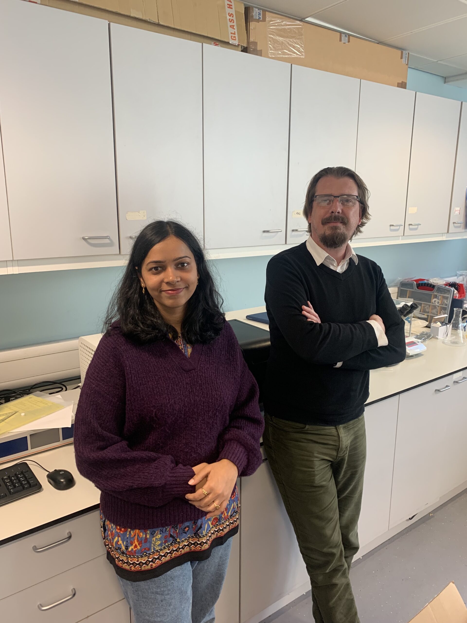 Sagarika (left) and Ross (right) they are stood in a lab in front of a workbench.
