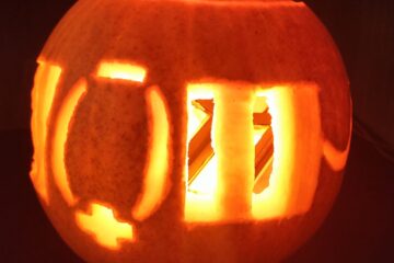 A pumpkin carved with parallel lines and a + and - symbol. Inside is one of the groups orange OLED lights.
