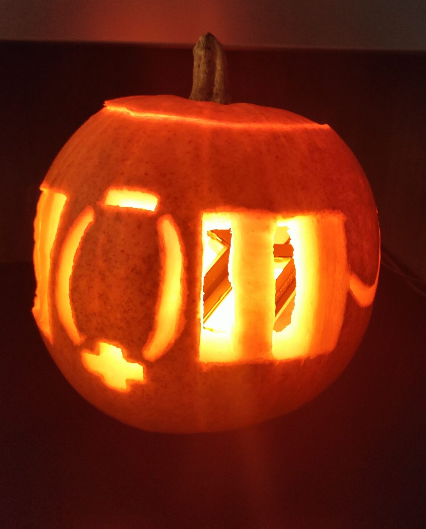 A pumpkin carved with parallel lines and a + and - symbol. Inside is one of the groups orange OLED lights.